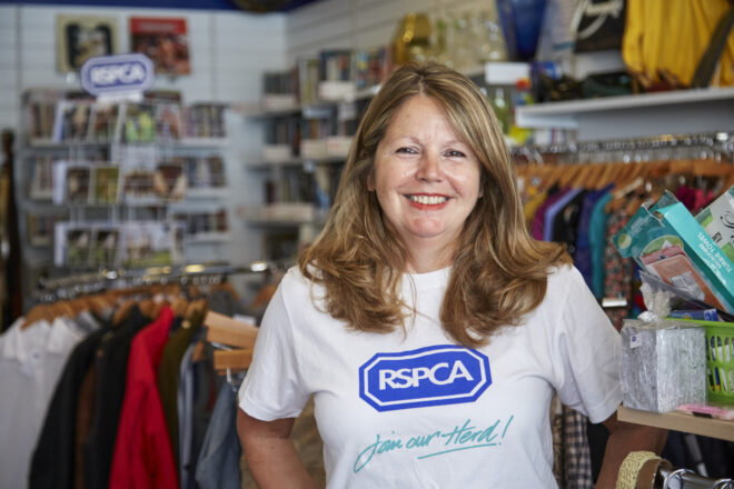 Lady working in RSPCA Charity shop, wearing RSPCA branded tshirt and similing at camera. Behind her are rails of donated clothes on hangers, shelves with books and other goods.